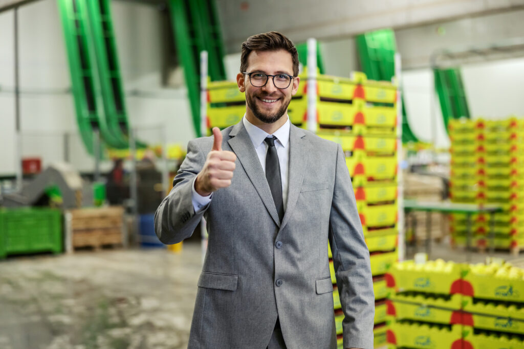 Approved by businessmen. A man dressed in style in a business gray suit stands in the warehouse and shows approval. Optimism during work and motivation of work staff. Thumbs up and smile, CEO manager